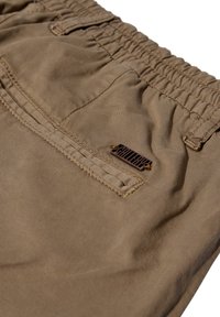 Brune elastiske buks med en baglomme og et lille rektangulært metaltag fastsat nær sømmen på lommen.