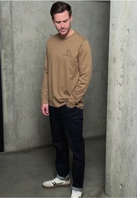 Dlouhý rukáv, světle hnědá košile s tištěným logem, spárovaná s tmavými džínami a bílými teniskami s hnědými akcenty. Stojící pozice u zdi.