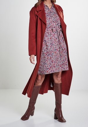 Femme portant un long manteau rouge par-dessus une robe à motifs longueur genou et des bottes marron à talons montantes jusqu'au genou, debout sur un fond blanc.