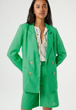 Groene linnen blazer met dubbelbreasted knopen, gecombineerd met bijpassende op maat gemaakte shorts en een witte geborduurde blouse eronder.