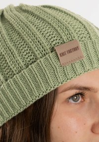 Berretto lavorato a maglia verde chiaro con motivo a coste e un'etichetta in pelle beige con la scritta "Knit Factory." Vista ravvicinata che mette in evidenza la texture e i dettagli.
