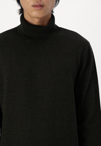 Młoda osoba ubrana w gładki czarny sweter z golfem, widoczna od ramion do nieco poniżej warg, na białym tle.