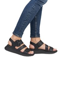 Sandali neri con cinturini, dotati di una suola testurizzata, caratterizzati da una fibbia ampia e cinturini regolabili, indossati con jeans blu aderenti.