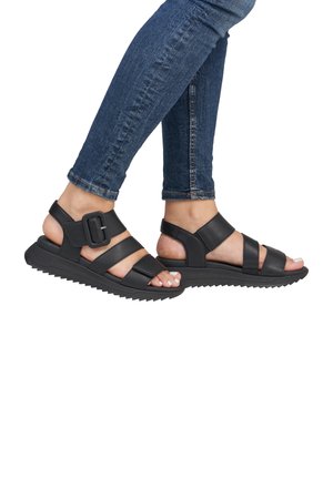 Sandalias negras con tiras y suela texturizada, con una hebilla ancha y correas ajustables, usadas con jeans ajustados azules.
