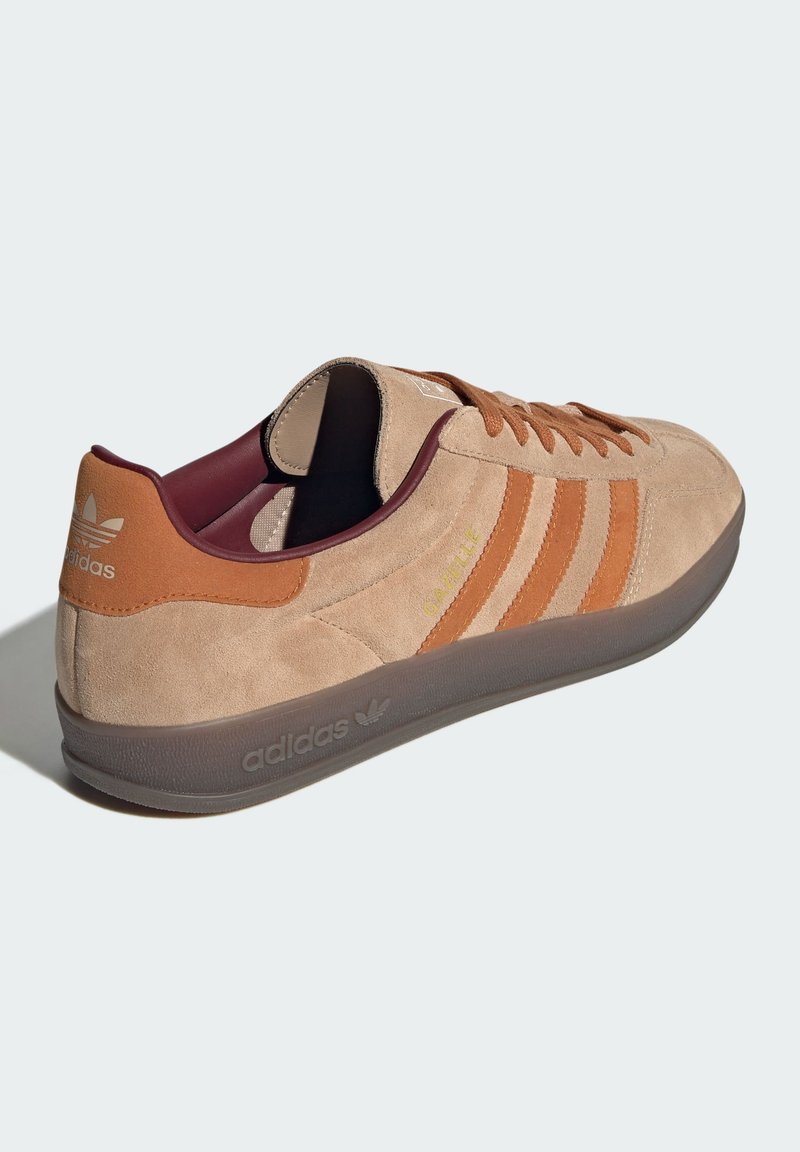 Zamszowe sneakersy w jasnym beżu z brązowymi akcentami. Posiadają trzy paski, wyściełany kołnierz oraz przezroczystą gumową podeszwę.