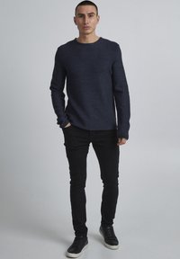 Dunkelblauer strukturierter Strickpullover mit rundem Ausschnitt, kombiniert mit schwarzen Slim-Fit-Jeans und schwarzen Sneakers mit weißen Sohlen.