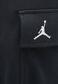 Μαύρο ύφασμα με μια μικρή ραμμένη τσέπη που διαθέτει μια λευκή κεντημένη σιλουέτα ενός παίκτη μπάσκετ που πηδά με μια μπάλα.