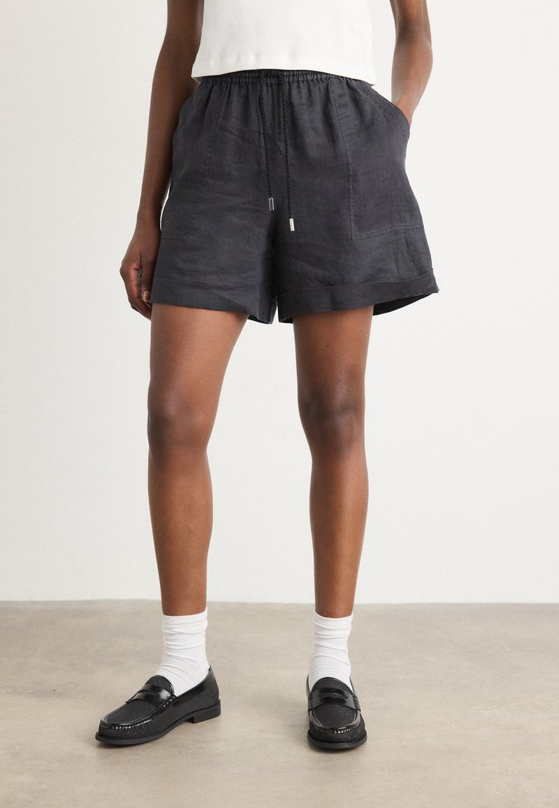 Pantaloni corti neri in lino con vita elastica e cordoncino, caratterizzati da tasche laterali e una vestibilità comoda, abbinati a mocassini neri e calze bianche.
