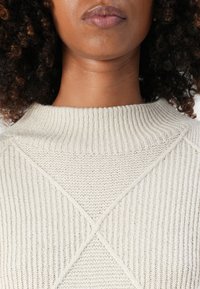 Hellbeige Pullover mit einem gerippten Kragen und einem diagonalen, strukturierten Muster über der Brust. Hergestellt aus einem weichen Strickmaterial.