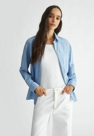 Camisa de botones a rayas azul y blanca con mangas largas, llevada sobre una camiseta de tirantes blanca de canalé, combinada con pantalones blancos.
