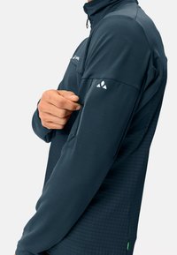 Donkerblauwe sportjack met een hoge kraag, ritszak op de arm en een gestructureerde stof. Bevat een contrasterend logo op de schouder.
