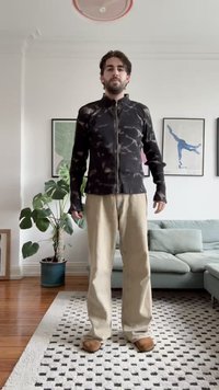 Svart hettejakke med marmor mønster, ribbet tekstur. Beige vide bukser, brune tøfler, står i et lyst rom med planter.