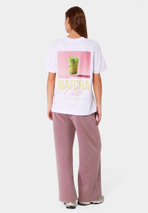 Kobieta ubrana w biały T-shirt z mrożonym napojem matcha i napisem „Matcha Club” na plecach, w zestawieniu z luźnymi liliowymi spodniami i trampkami.