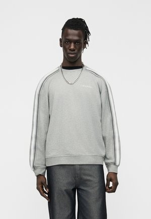 Muž s tmavou pokožkou a opletenými vlasmi, ktorý má na sebe sivú mikinu Calvin Klein s pruhovanými rukávmi a tmavé džínsy, stojaci pred bielym pozadím.