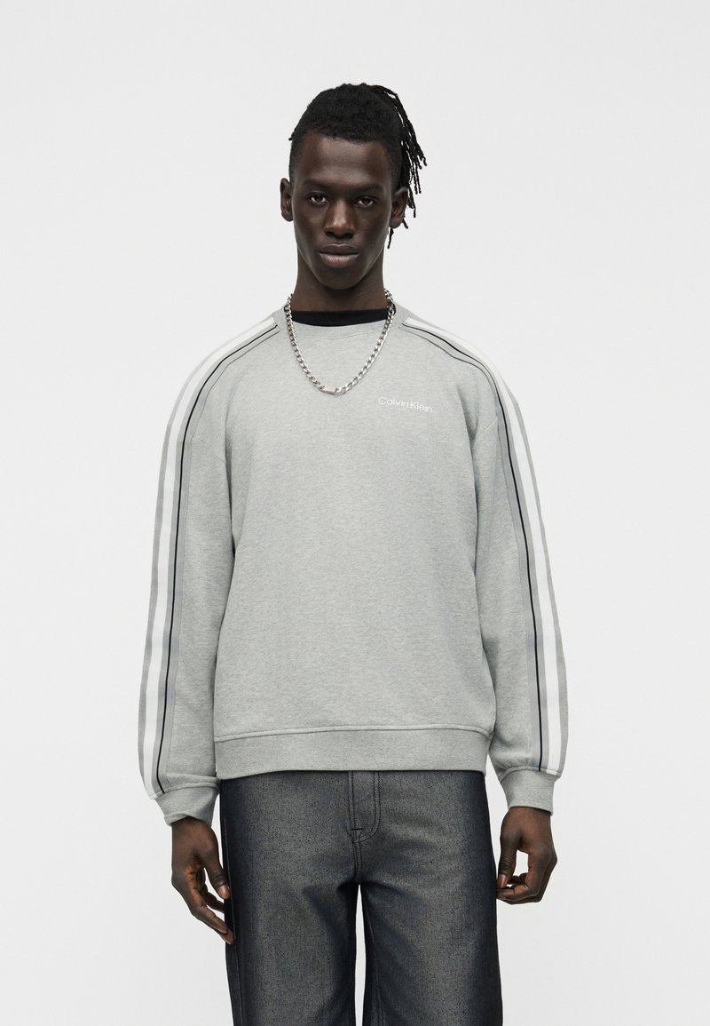 Άνδρας με σκουρόχρωμο δέρμα και πλεξούδες, που φοράει γκρι φούτερ Calvin Klein με ριγέ μανίκια και σκούρο τζιν, στέκεται μπροστά σε λευκό φόντο.