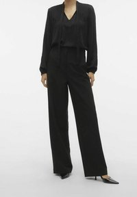 Blouse noire avec détails plissés et liens, associée à un pantalon noir taille haute à jambes larges. Le mannequin porte des escarpins noirs à bouts pointus.