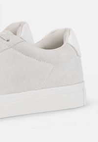 Light grey suede sneaker with a smooth white heel counter, rounded toe, and flat white rubber sole. Detailed stitching along the upper.