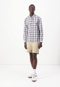 Checked button-up shirt in white, red, and navy; khaki shorts; white socks; beige sneakers. Model standing against a plain background.