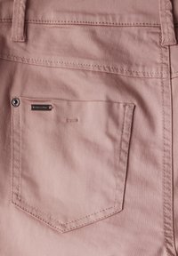 Pölyinen vaaleanpunainen puuvillakangas, jossa on takatasku, jossa on hopeanvärinen nappi ja ommeltu yksityiskohta, esittelee sileää materiaalia ja siistejä linjoja.