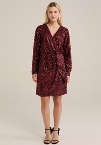 Rode pailletten jurk met een wikkeldesign, lange mouwen, een voorste twistdetail en een korte zoom. Gepaard met zwarte bandjeshakken.