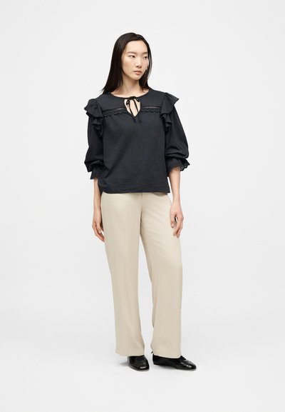 Femme debout vêtue d'un chemisier noir à volants avec un col à nouer, un pantalon beige à jambes larges et des chaussures noires sur un fond blanc uni.