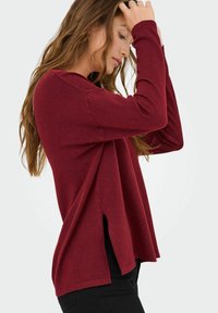 Maglione lungo bordeaux a maniche lunghe con una vestibilità comoda, presenta spacco laterale, una texture morbida e un collo tondo. Abbinato a pantaloni neri.