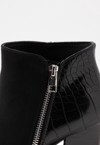 Sorte ankelstøvler med sidelukning, tekstureret krokodillemønster på hælområdet og glat ruskindslignende materiale foran.