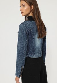 Chaqueta de denim en azul oscuro con un acabado desgastado. Presenta un diseño corto, puños con cremallera y cuello clásico.