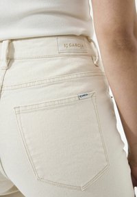 Roomkleurige spijkerbroek met een hoge tailleband, voorzien van een achterzak en een merkpatch. Glad denimtextuur met subtiele stikselaccenten.