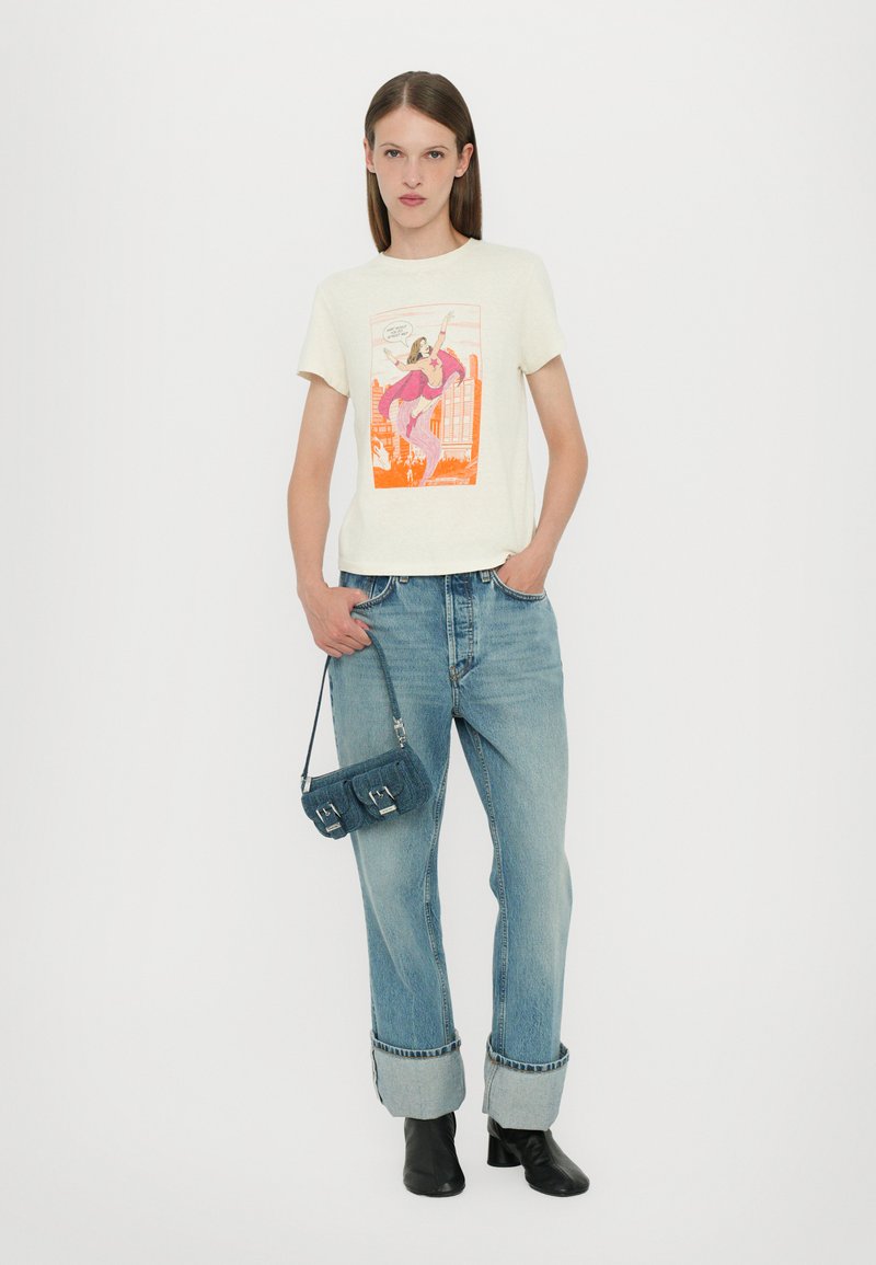 Cremfarbenes T-Shirt mit einem bunten Grafikdesign, kombiniert mit hellblauen lässigen Jeans. Das Model hält eine kleine dunkelblaue Handtasche und trägt schwarze Stiefel.