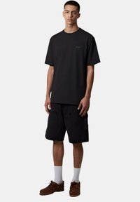 T-shirt in cotone nero con scollatura rotonda e maniche corte, abbinato a pantaloncini di jeans neri. Il modello indossa scarpe marroni e calzini bianchi.