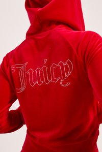 Červená velúrová mikina s kapucňou a veľkým logom "Juicy" posiatym kryštálmi na chrbte. Má klokanka a rebrované manžety.
