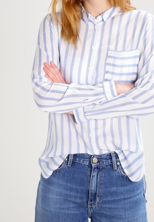 Osoba nosící světle modrou a bílou košili s vertikálními pruhy a náprsní kapsou, se zkříženýma rukama, v kombinaci s modrými džíny.