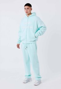 Felpa con zip verde menta e pantaloni della tuta abbinati, realizzati in tessuto morbido. Presenta tasche, logo bianco sul petto e vita con cordino.