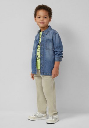 Junger Junge steht, trägt eine Jeansjacke über einem grünen T-Shirt mit Grafik, beiger Hose und weißen Turnschuhen vor einem einfarbigen Hintergrund.