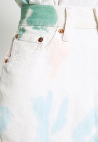 Primer plano de unos vaqueros de mezclilla blancos con remaches de cobre y patrones de tie-dye en tonos verde pastel, azul y durazno cerca del bolsillo delantero y la cinturilla.