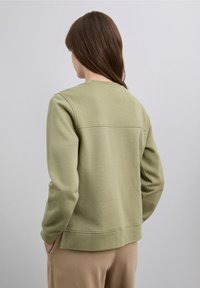 Zielona bluza wykonana z teksturowanej tkaniny, z okrągłym dekoltem i długimi rękawami. Na plecach znajduje się poziomy szew ozdobny.