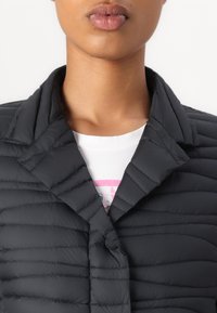 Casaco preto matelassado com colarinho, apresentando um fecho com botão e detalhe de costura horizontal. Por baixo, uma t-shirt branca é visível.