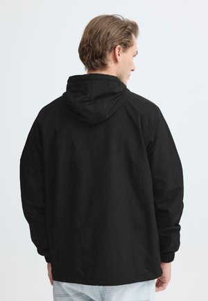 Man die een zwarte capuchonjas draagt, gezien van achteren tegen een effen lichte achtergrond.