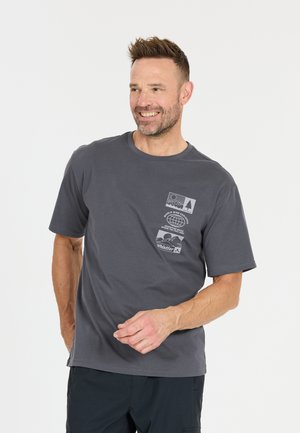 Lächelnder Mann mit kurzen Haaren, der ein dunkelgraues T-Shirt trägt, auf der linken Brust mit drei grafischen Motiven zum Thema Outdoor.