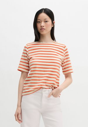 Kobieta z długimi czarnymi włosami, ubrana w pomarańczowo-biały pasiasty krótki rękaw i jasne spodnie, stoi z jedną ręką w kieszeni.