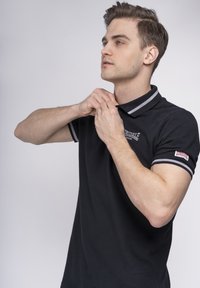 Camisa polo negra con cuello y puños a rayas grises, con un parche de logo en la manga izquierda. El modelo ajusta el cuello.