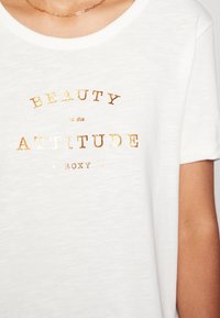 Weißes T-Shirt mit kurzen Ärmeln aus weichem Stoff, mit metallic-goldener Aufschrift "SCHÖNHEIT ist eine EINSTELLUNG" und dem Markennamen "ROXY" darunter.