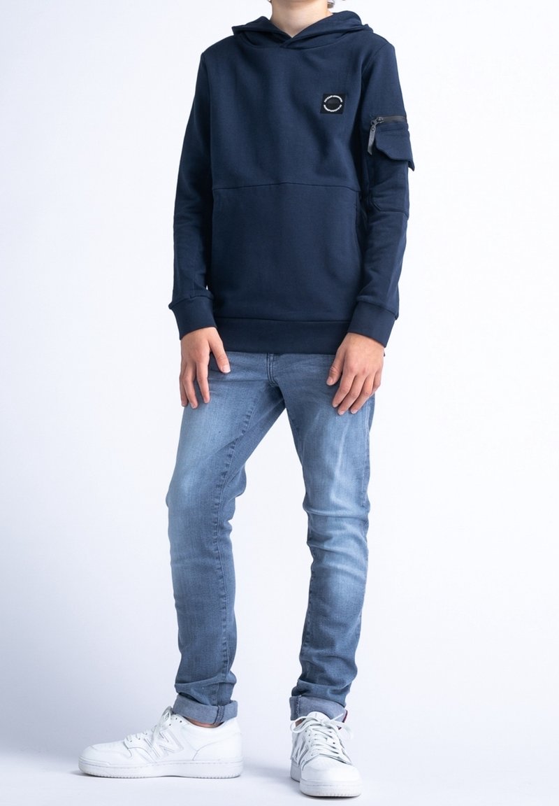 Sweat à capuche bleu marine avec poche zippée sur la manche, tissu texturé et poignets côtelés, associé à un jean bleu clair et des baskets blanches.