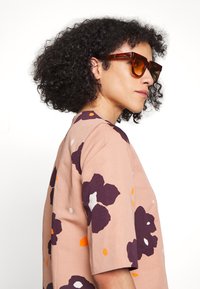 Blusa rosa com grande padrão floral em roxo escuro e detalhes em laranja. Modelo usa óculos de sol castanhos. Mangas curtas e decote redondo.