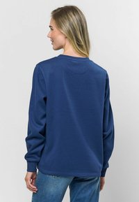 Tumši zila garām piedurknēm brīvais krekls ar apaļu kakla izgriezumu, mīksts tekstūras materiāls un ribotas piedurknes. Aizmugurē ir smalka šuvju detaļa.