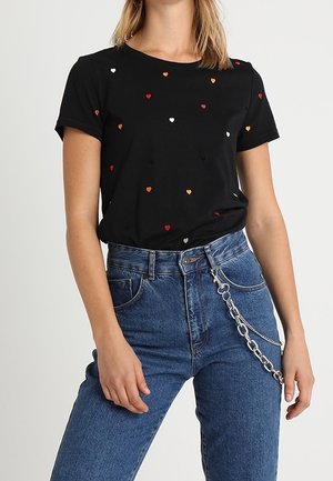 Czarny t-shirt z krótkim rękawem z małymi czerwonymi, pomarańczowymi i białymi wzorami serc, połączony z niebieskimi wysokim stanem jeansami i srebrnym łańcuszkiem jako dodatkiem.