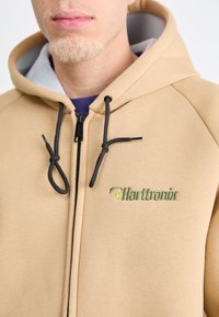 Tan zip-up jopica iz mehkega blaga, s sivim notranjim kapuco in črnimi vrvicami, ter z zelenim vezenim logotipom na prsih.