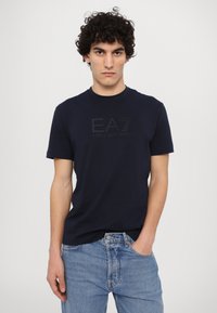 Ναυτικό μπλε βαμβακερό μπ λουζάκι με κοντά μανίκια, που διαθέτει ανάγλυφο λογότυπο EA7 σε μαύρο χρώμα και στρογγυλή λαιμόκοψη, συνδυασμένο με ανοιχτό μπλε τζιν.