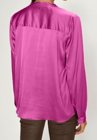 Blouse rose satinée à manches longues, avec un bouton au col et une texture lisse. Le dos présente un design simple sans motifs.